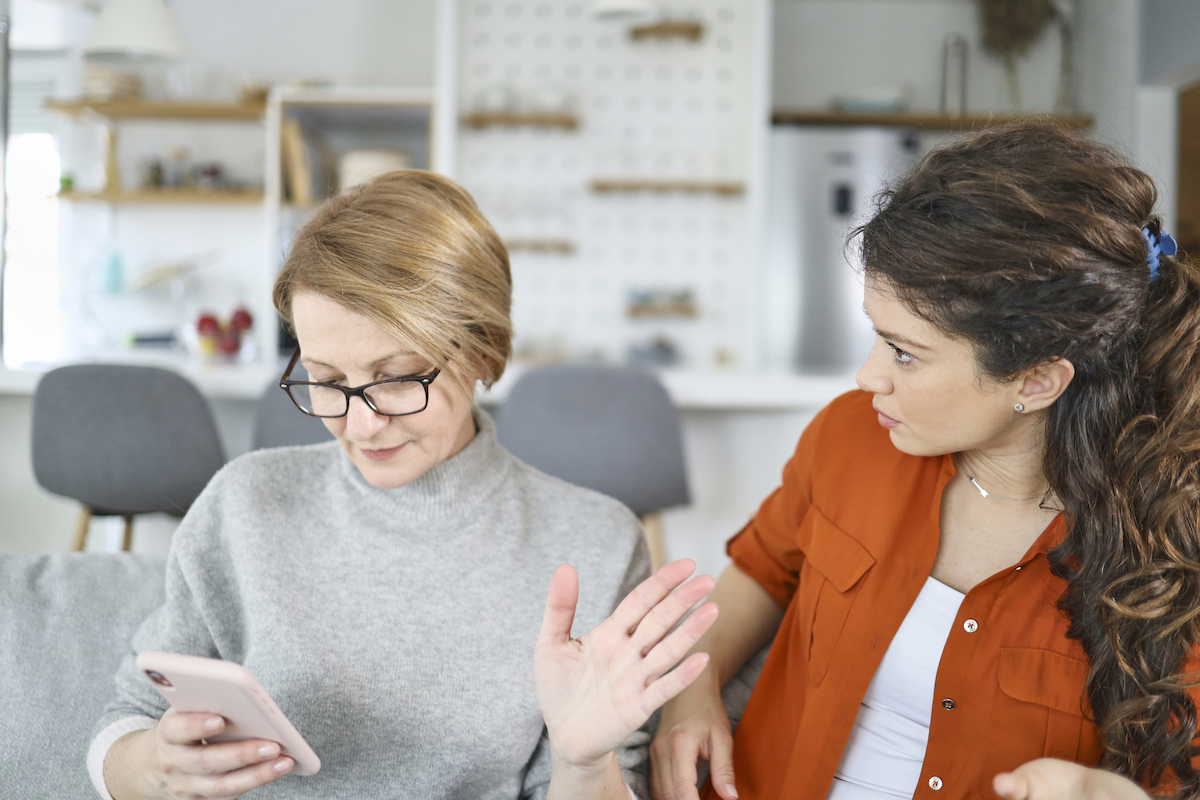 Shot of a senior mother looking at the phone while upset daughter looks at her on the couch.