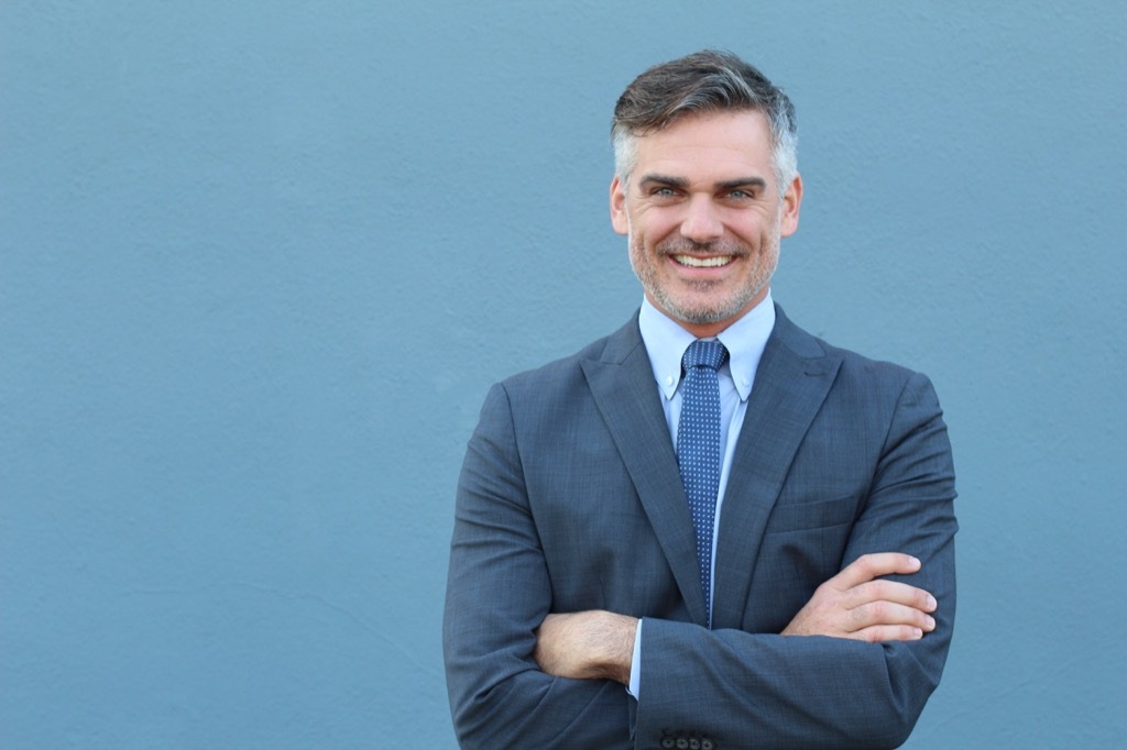 man with grey hair and blue eyes stands with crossed arms in suit on blue background