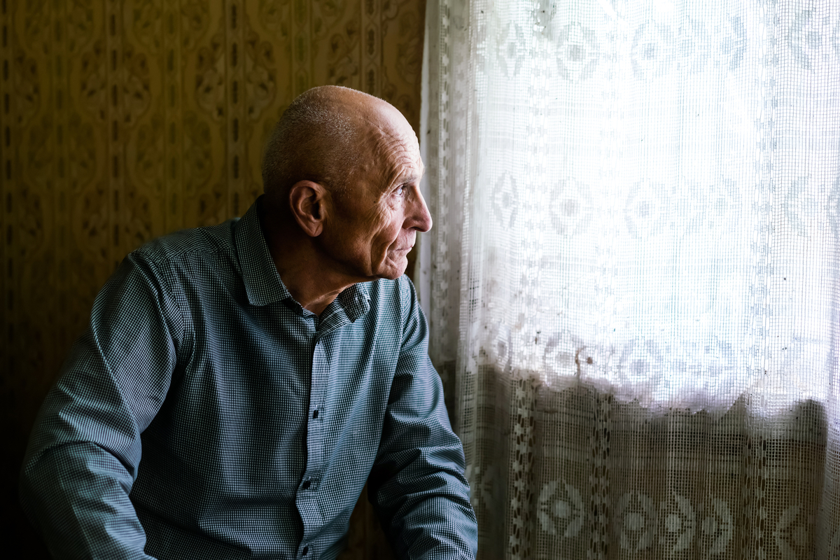 older white man looking out window feeling apathetic