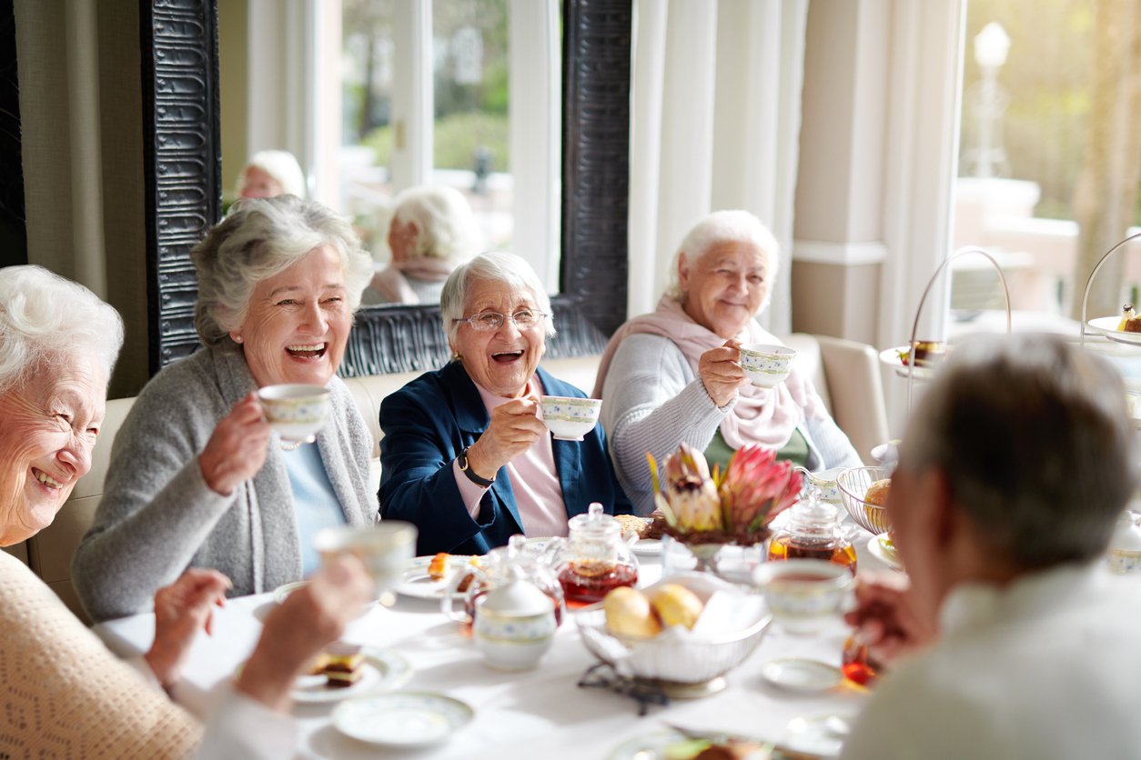 A group of senior women sitting around a table eating and drinking tea while smiling