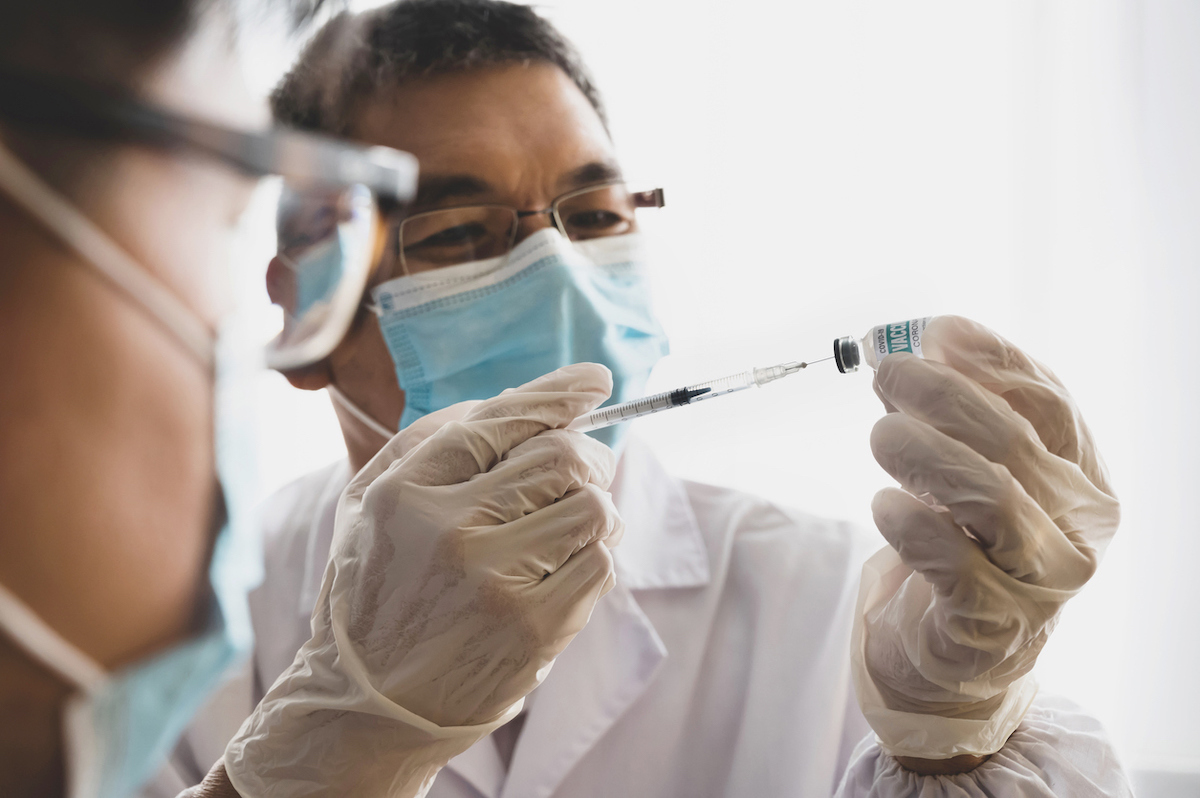 Doctor in protective face mask wearing surgical gloves injecting vaccine to a patient in medical lab.