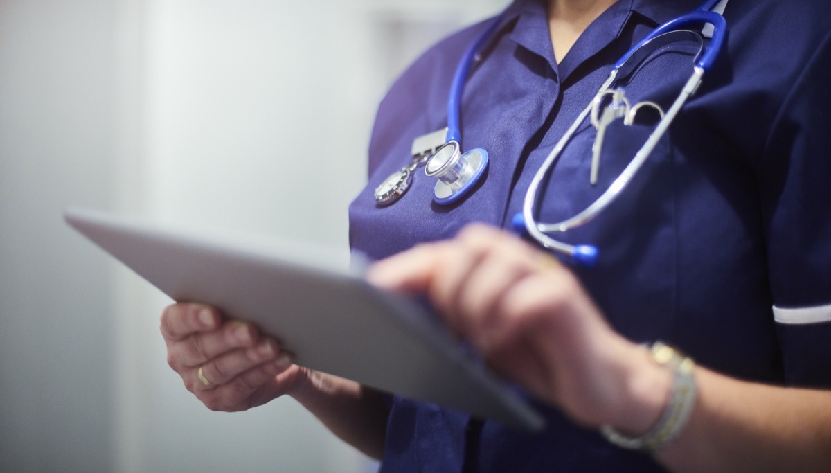 female surgeon typing on digital tablet in hospital or surgery. She is wearing a dark blue nurse‚Äôs top and has her stethoscope around her neck. She is looking at her patients records on her digital tablet and sending emails.