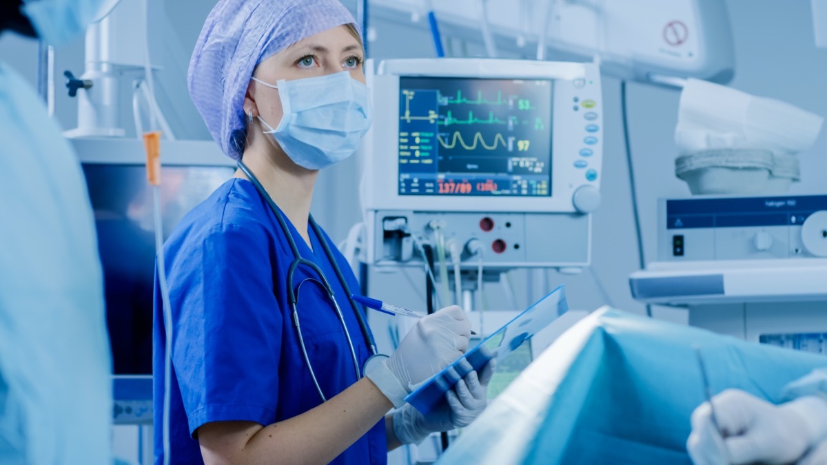 In the Hospital Operating Room Anesthesiologist Looks and Monitors and Controls Patient's Vital Signs, Nodding to a Chief Surgeon to Proceed with Surgery.