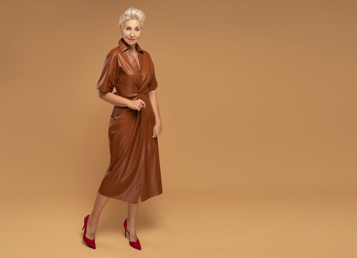 A mature woman with short blonde hair wearing a brown leather dress and red heels against a light brown background.