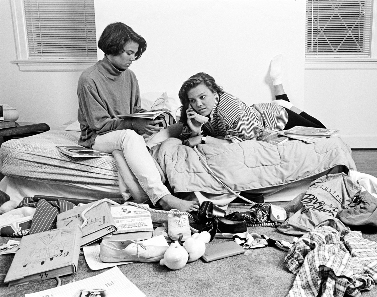  TWO TEENAGE AFRICAN AMERICAN GIRLS IN CLUTTERED BEDROOM ONE TALKING ON THE PHONE ONE READING MAGAZINE