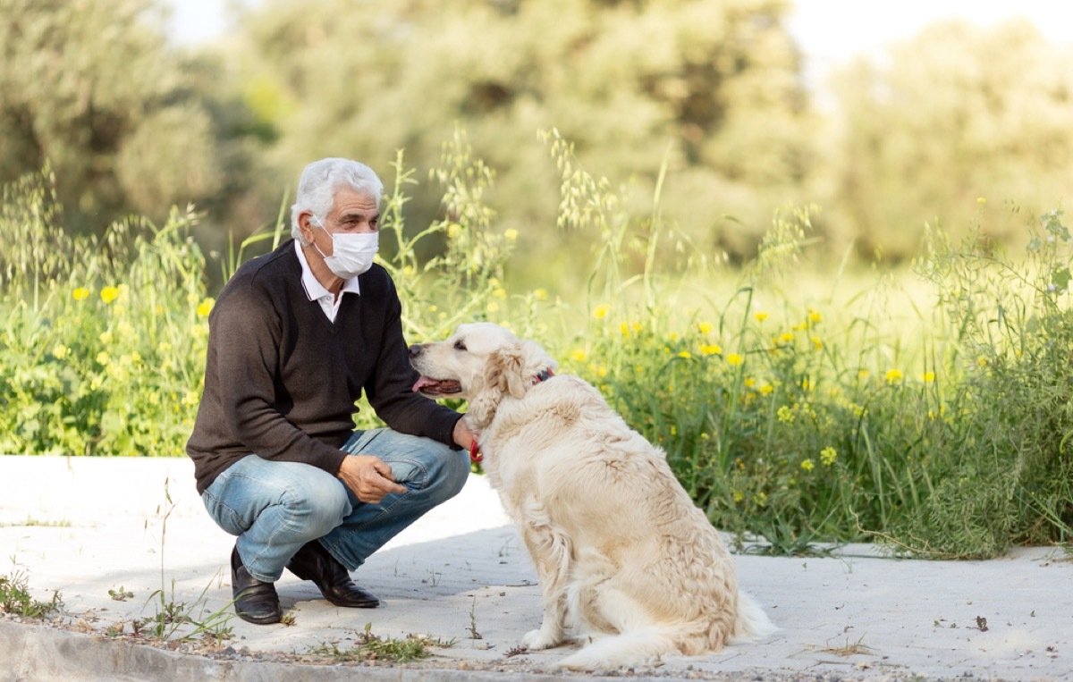 older white man with a face mask crouching down next to his dog outside