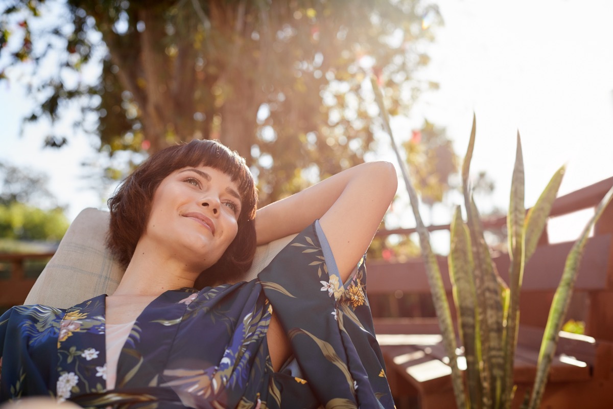 Young woman looking deep in thought and smiling while lying back in a deck chair on her patio on a sunny afternoon