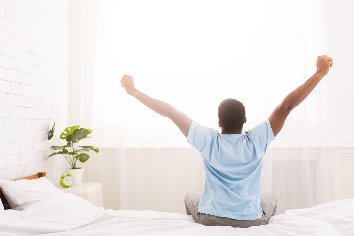 Black man pictured from behind stretching in bed in the morning, wearing a blue shirt benefits doing nothing