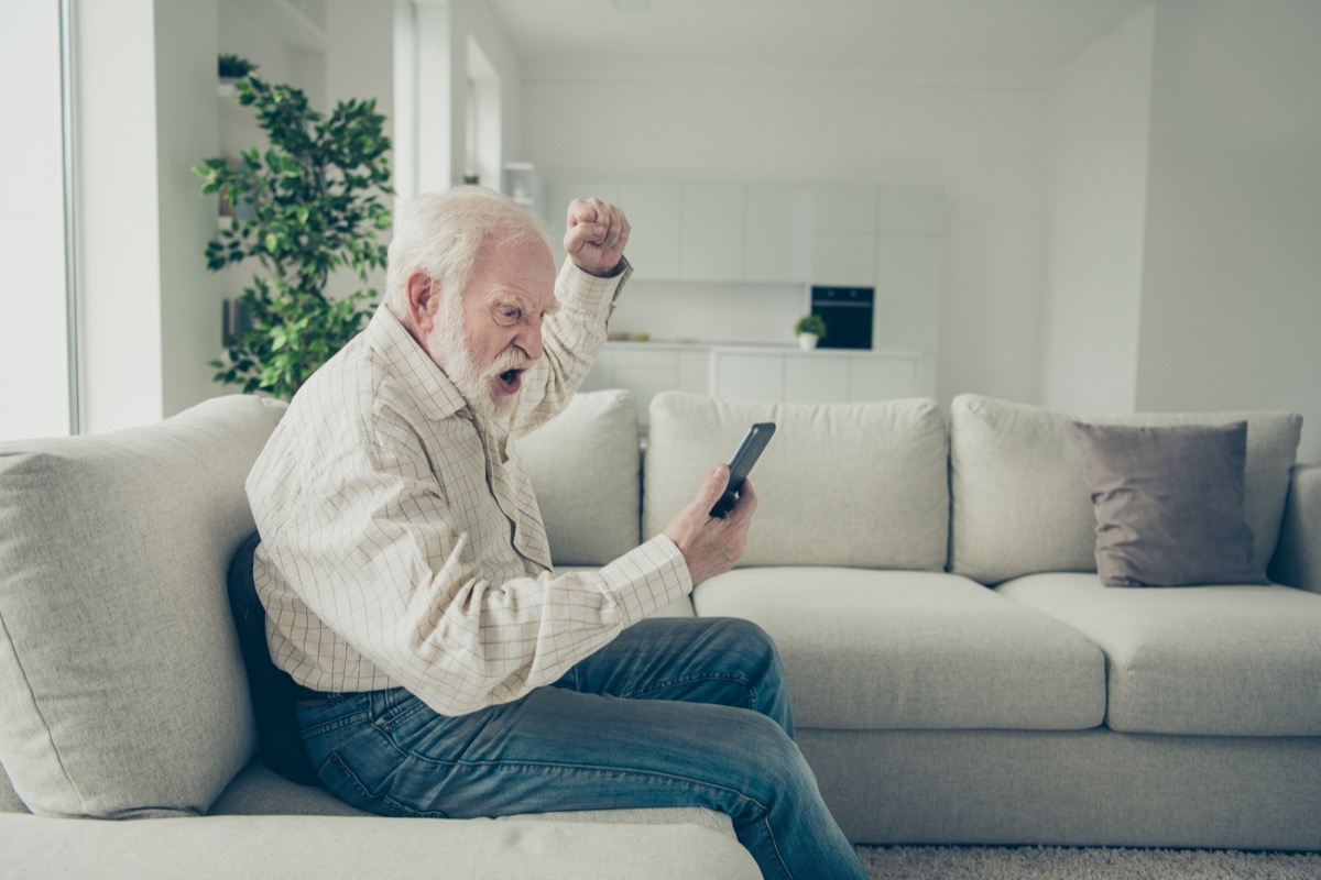 Profile side view portrait of mad stylish old man wearing checked shirt sitting on divan holding in hand phone going crazy resenting in white light modern interior studio (Profile side view portrait of mad stylish old man wearing checked shirt sitting