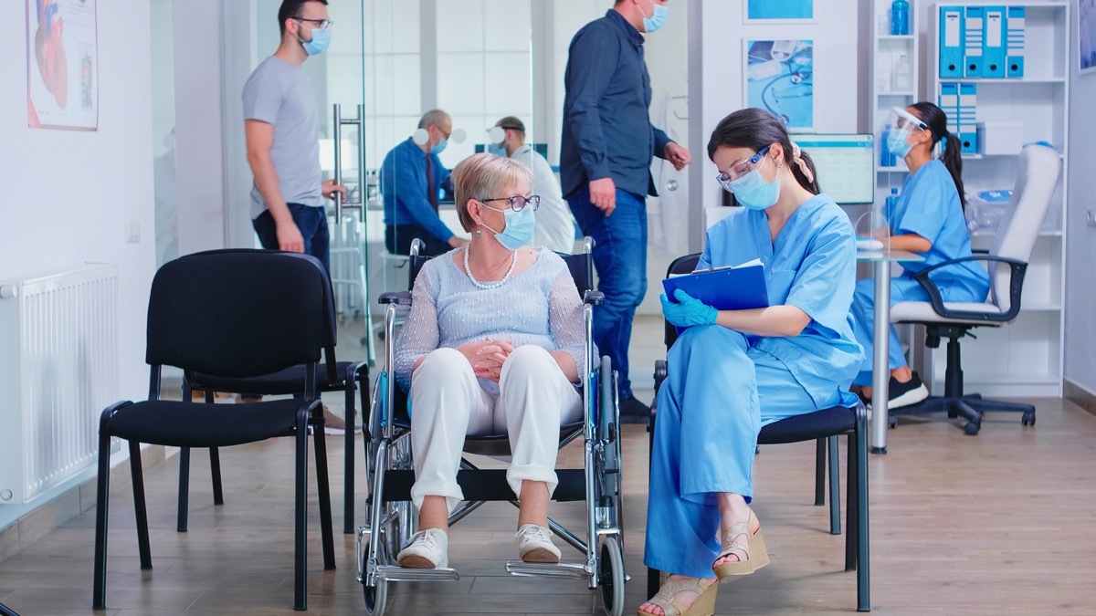 Nurse wearing face mask against coronavirus taking notes on clipboard while talking with disabled senior woman in wheelchair