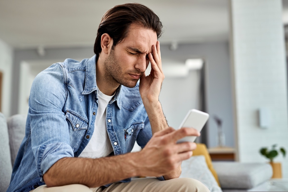 man using smart phone and holding his head in pain at home