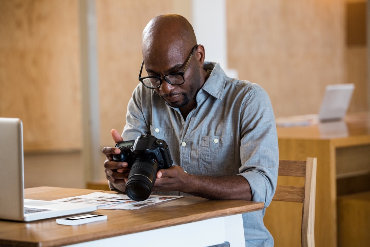 Black man in denim shirt with black glasses holds camera in front of computer, downsizing your home