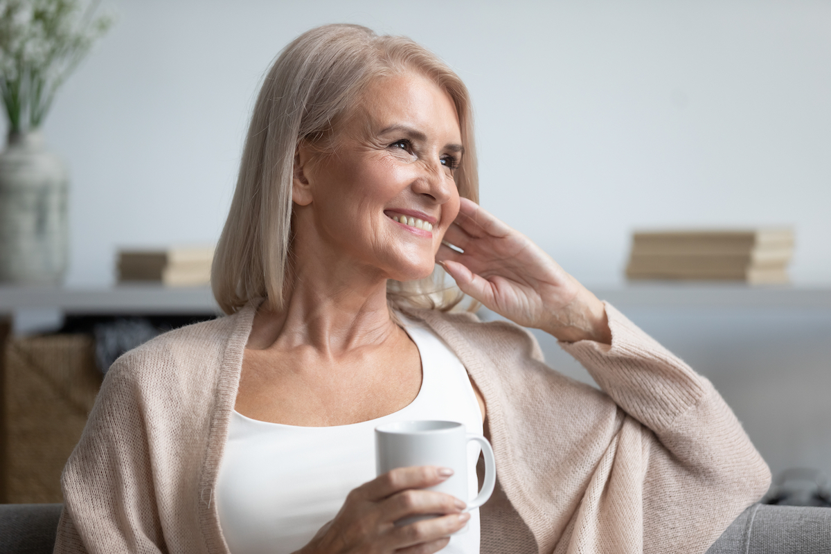 A happy mature woman sitting on her couch with a cup of coffee, gazing to the side.