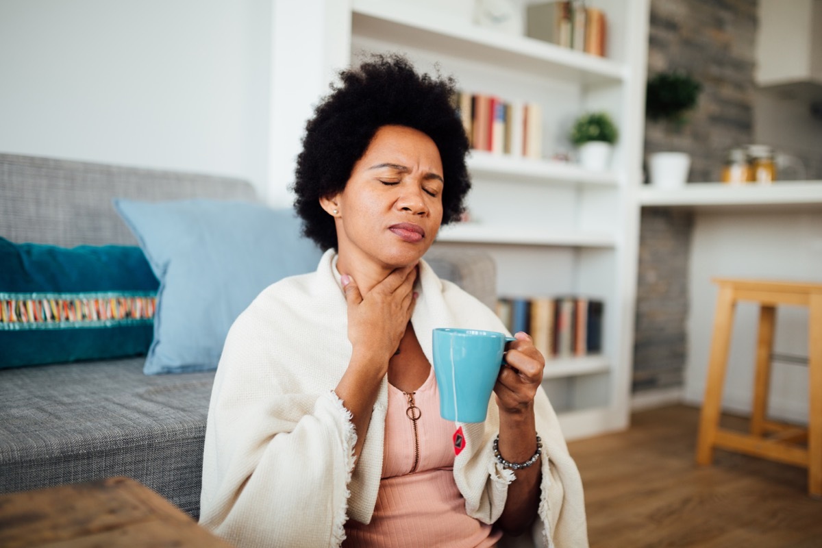 woman, fallen ill is staying at home wrapped in a blanket socially distancing and quarantining herself, feeling her throat hurt and being sore, having a cup of hot tea