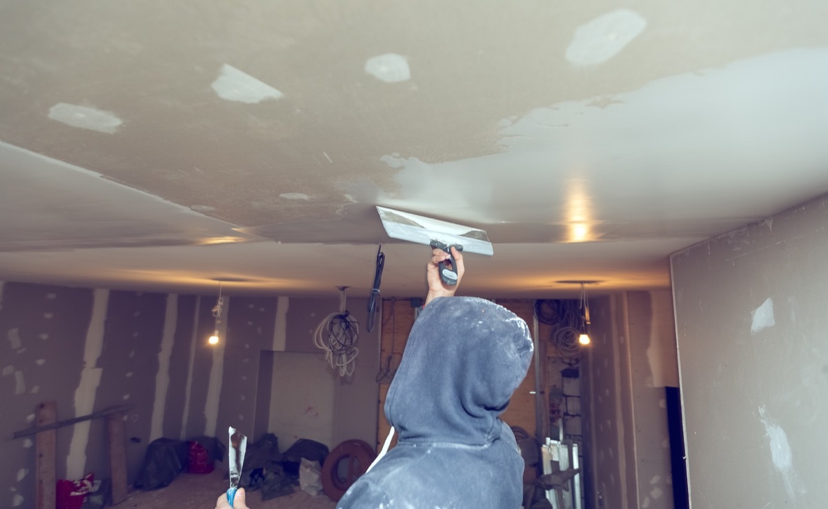 person in hoodie skim coating a drywall ceiling in unfinished room