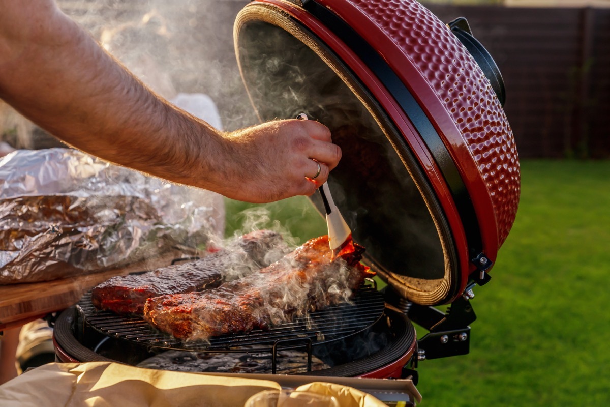 Smoke coming out of red ceramic Barbecue Grill. The man coats pork ribs with barbeque sauce. Picnic in modern homes terrace.