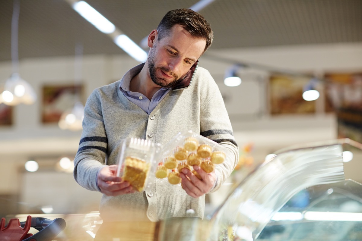 Portrait of confused middle-aged man doing grocery shopping in supermarket: calling his wife asking what dessert to choose