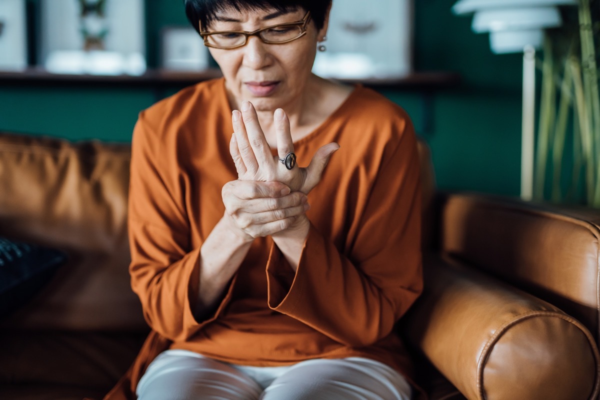 woman rubbing her hands in discomfort, suffering from arthritis in her hand while sitting on sofa at home. Elderly and health issues concept