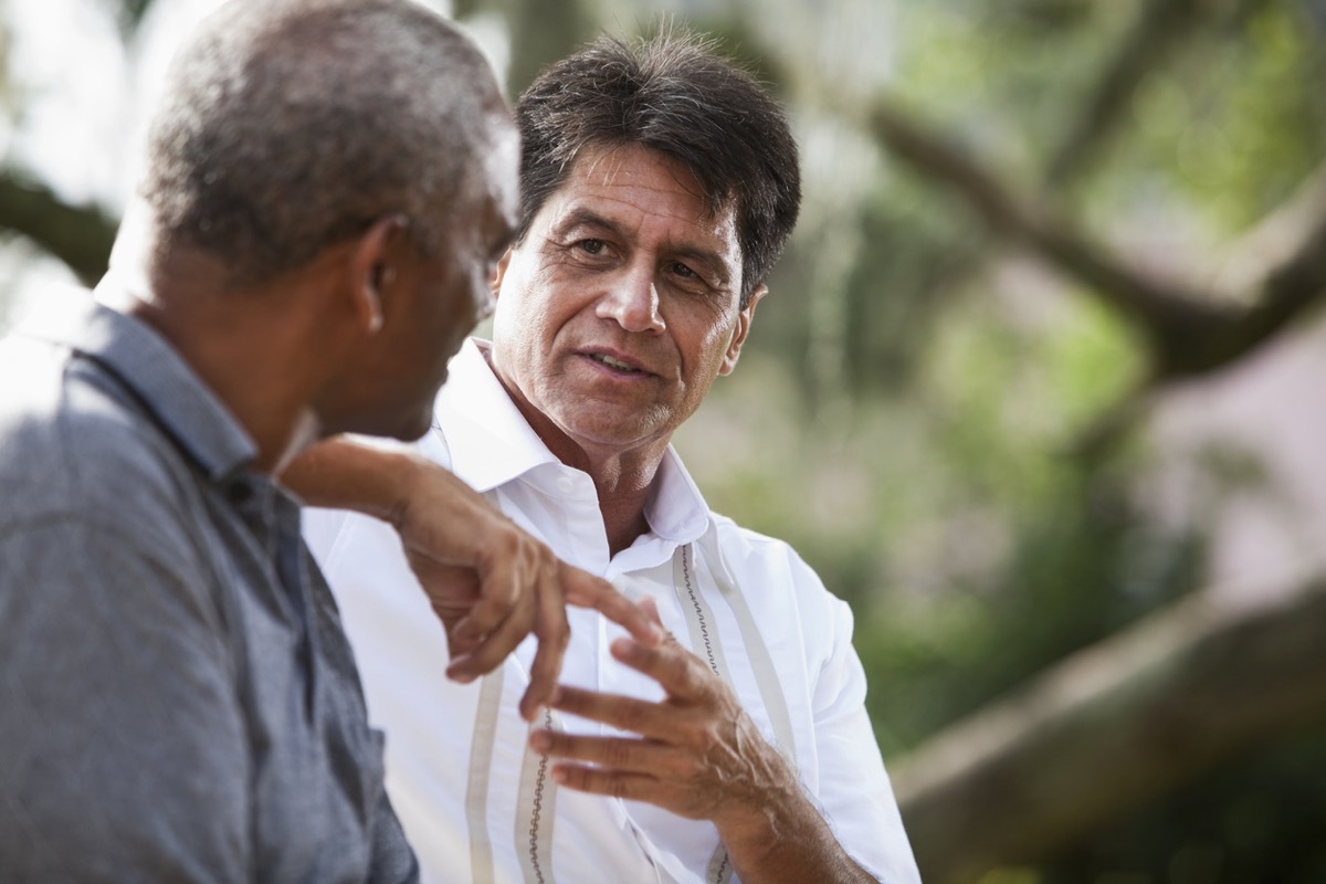 men (50s and 60s) sitting outdoors, having conversation. 