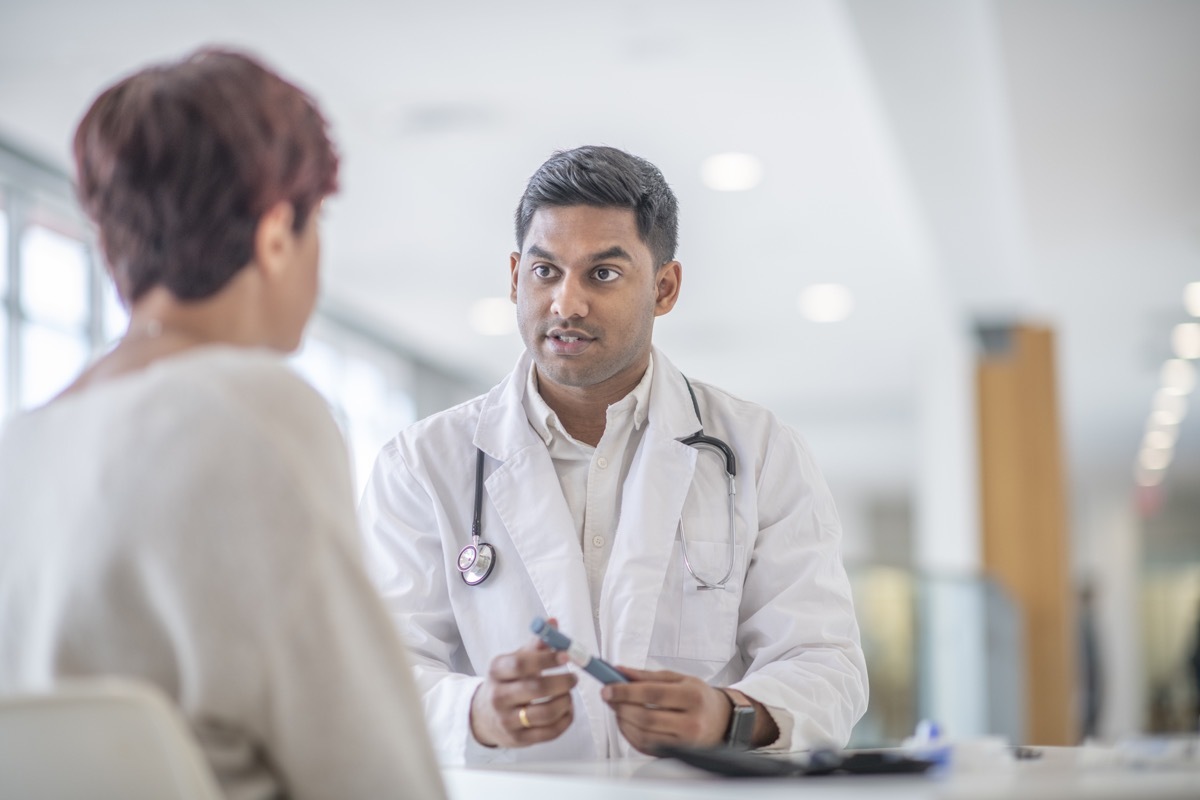 A female adult is at a check up appointment for a diabetic medical condition. She is sitting across the table from her Indian doctor who is holding a glucometer pen that is used to test her blood sugar levels.