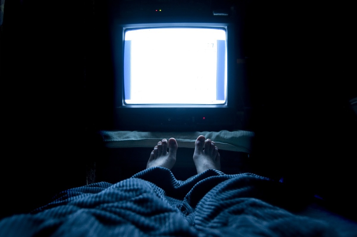 Person watches TV at night in his bed with his feet sticking up out of the blankets. The TV screen is blank white