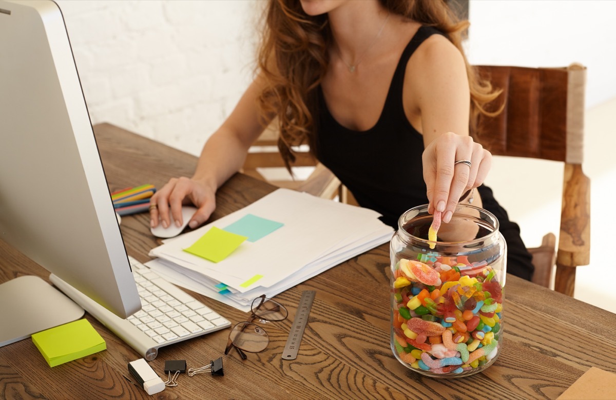 Woman mindlessly eating a jar of gummy candy while working on her desktop computer