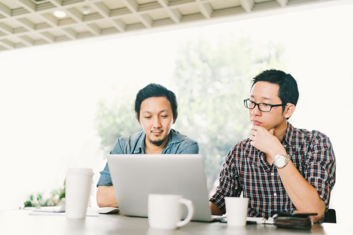 two male business colleagues or college students work together using laptop, startup project meeting or teamwork brainstorm concept, at coffee shop or modern office