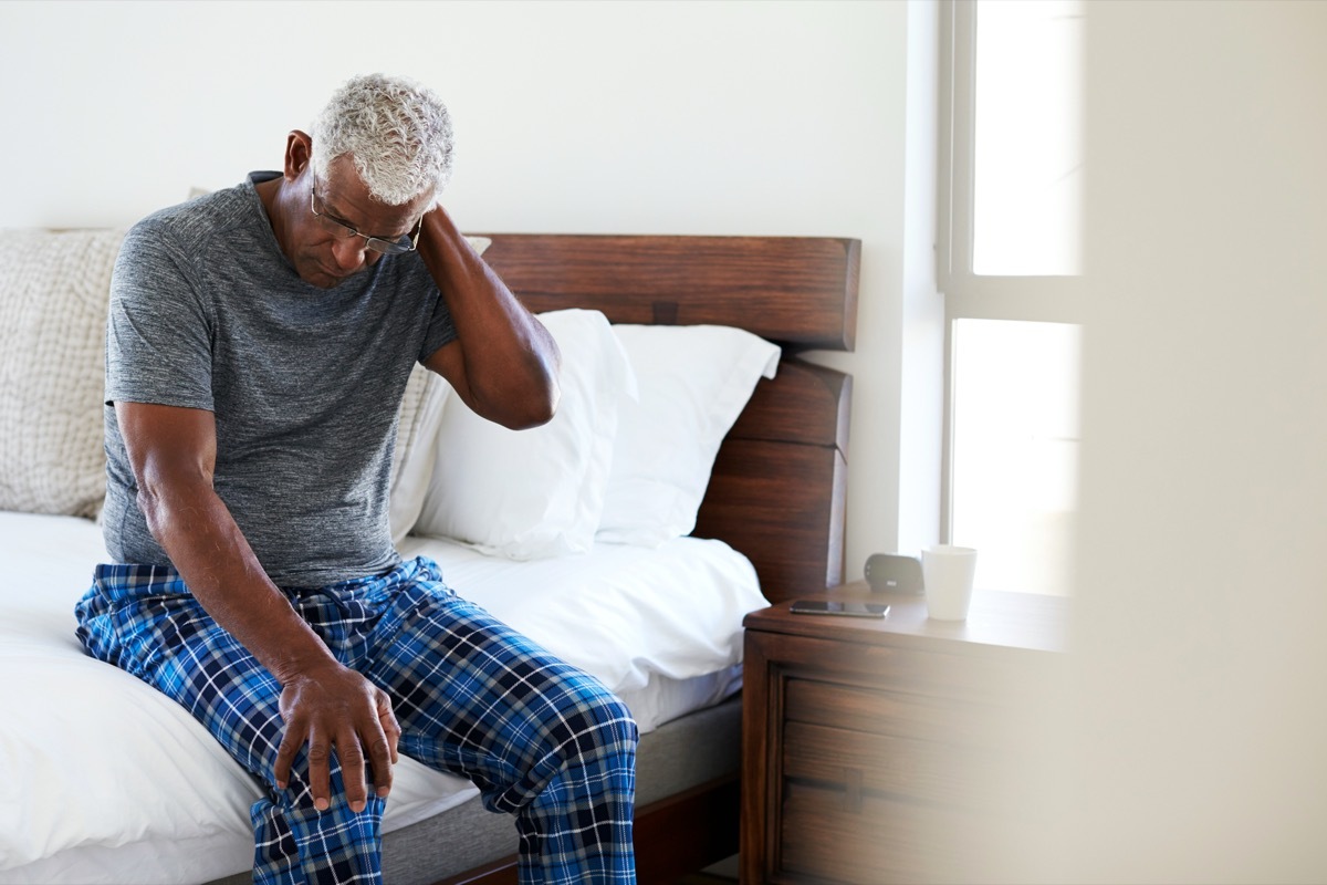 Senior Man Suffering With Neck Pain Sitting On Side Of Bed At Home