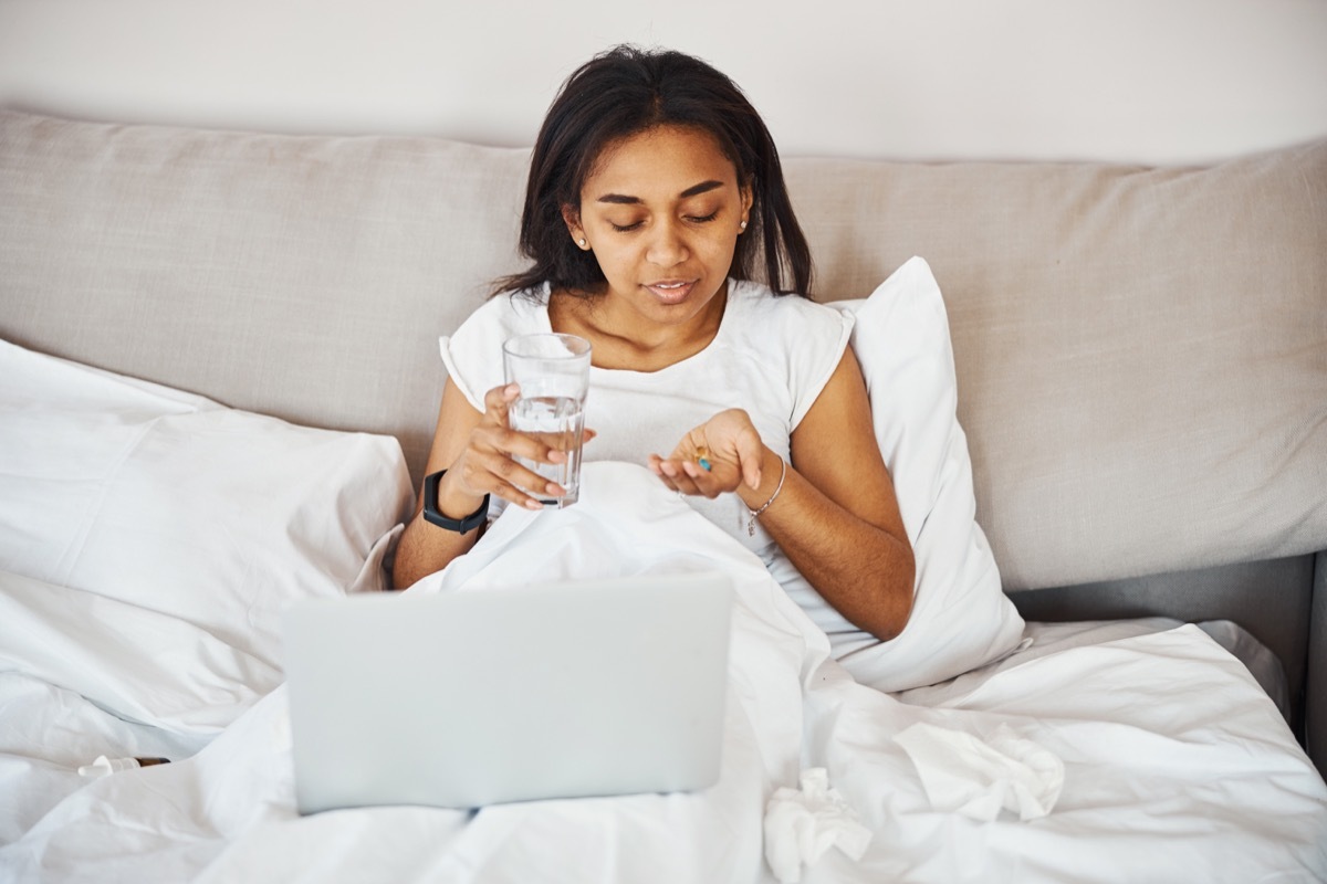 lady lying in bed with laptop and smiling while holding pill and glass of water