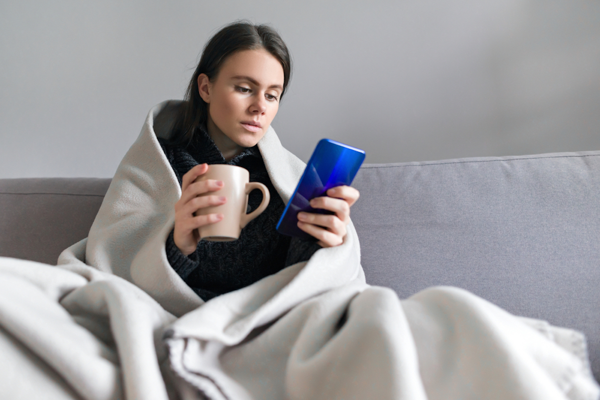 young woman wrapped in blanket with mug, looking at phone