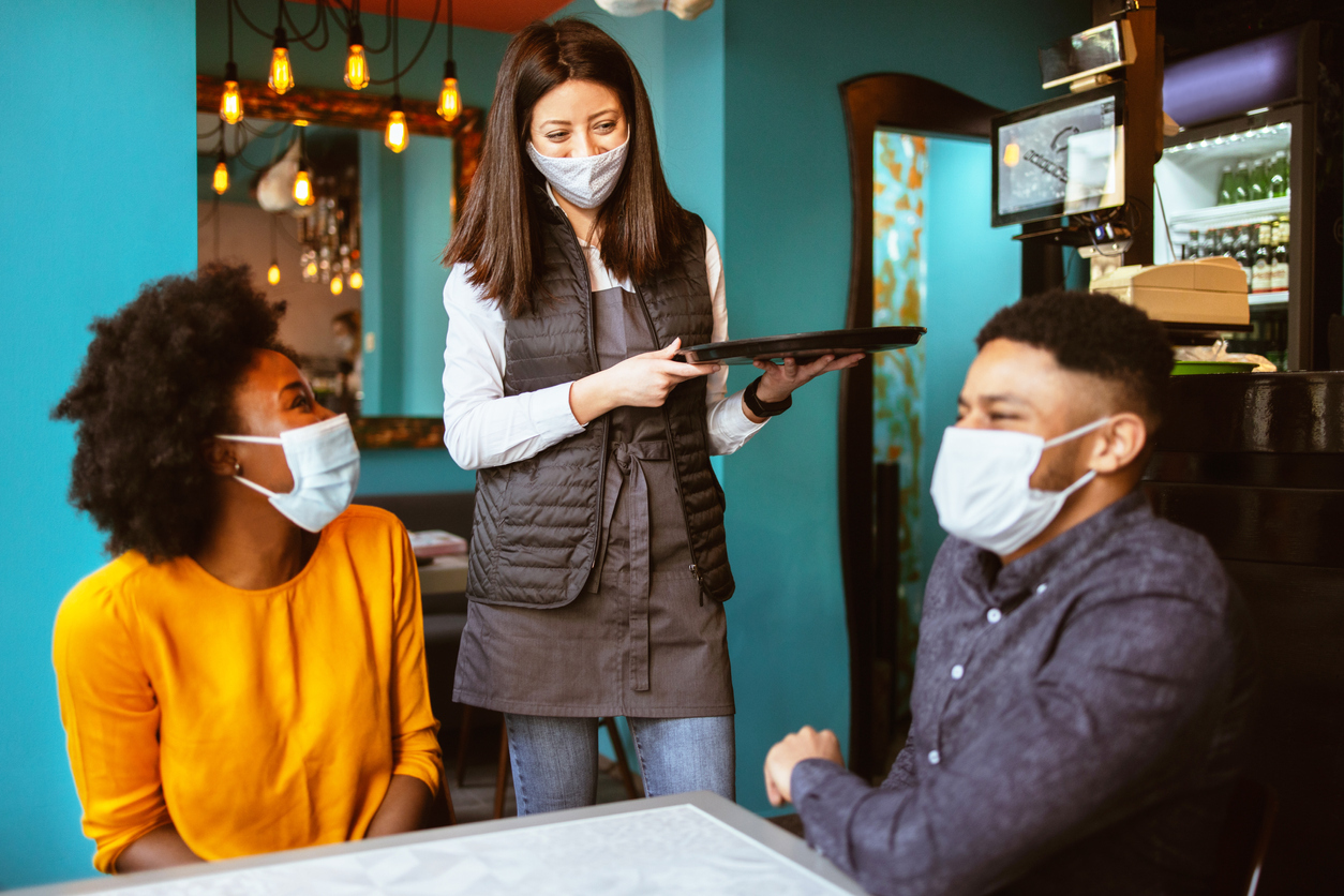 A young server speaks to a couple sitting at a table at a restaurant while all wear face masks