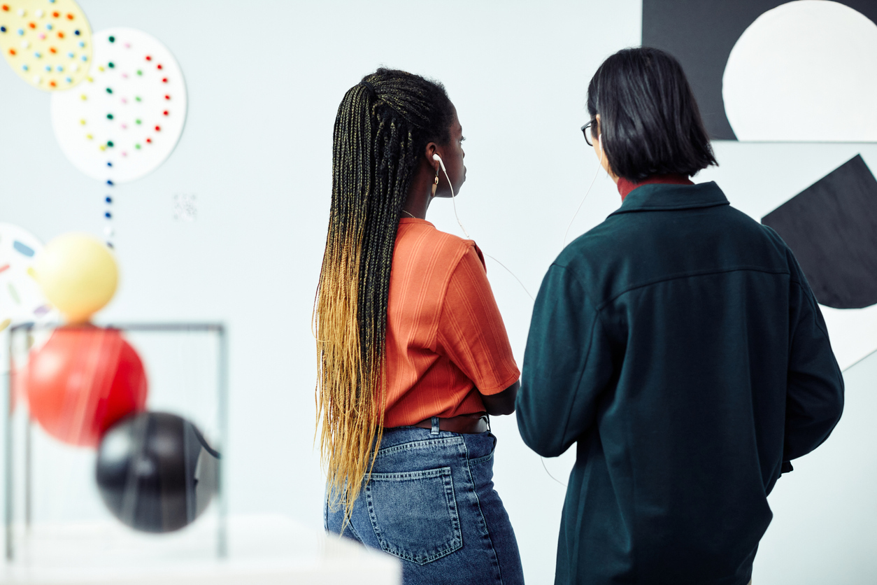 Rear view of ethnically diverse young man and woman looking at abstract painting and listening to audio tour at exhibition in art gallery