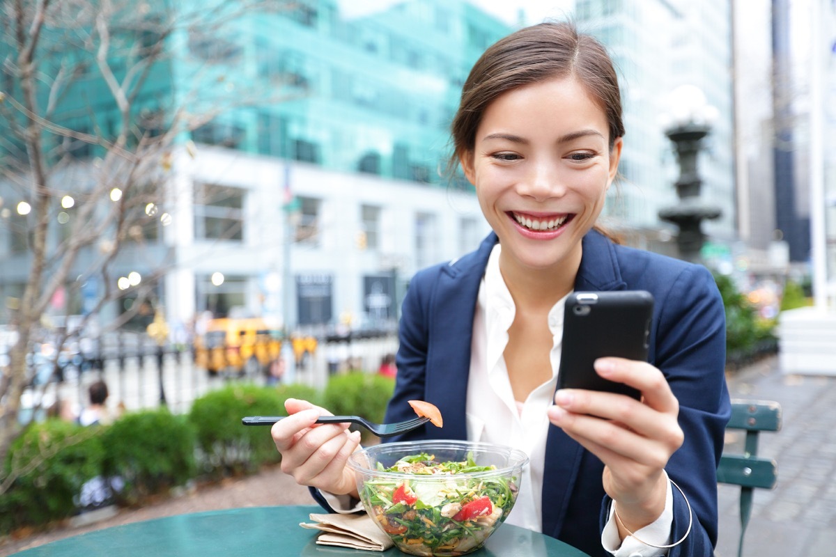 Woman smiling while eating a salad for lunch alone looking at her phone