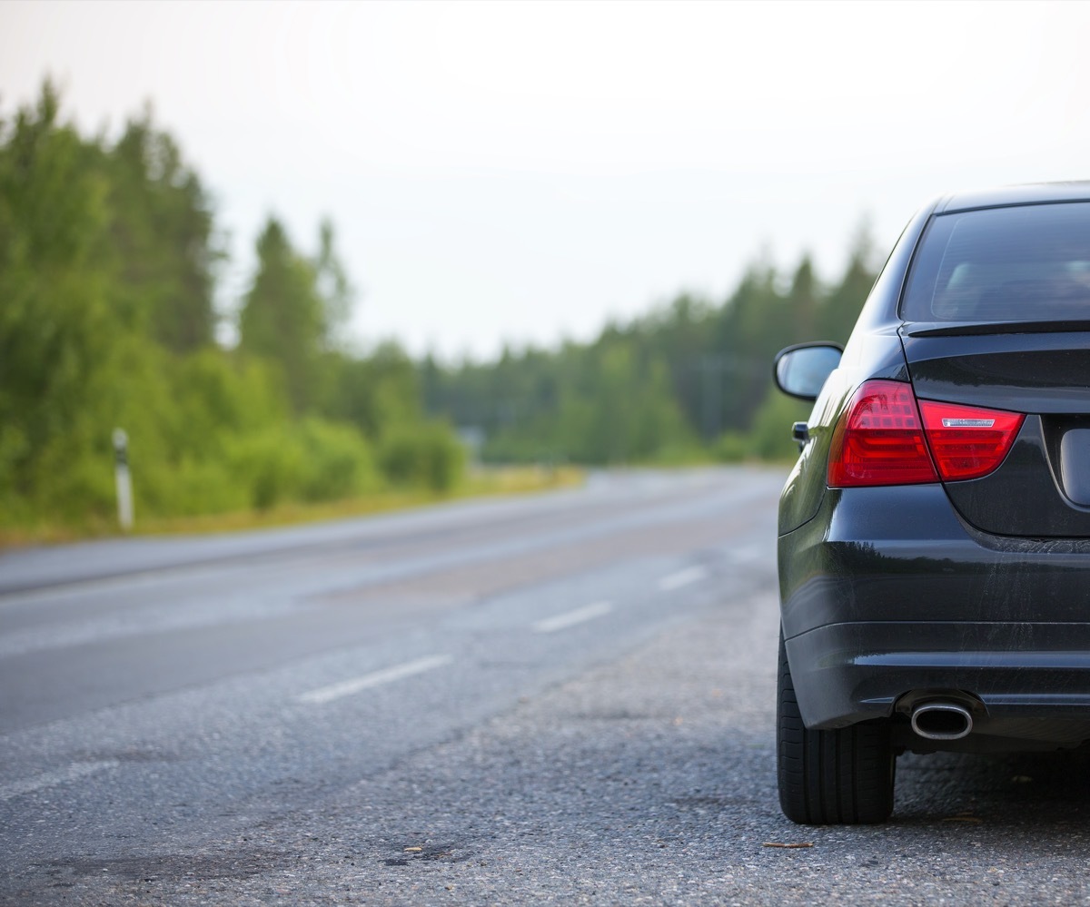 A black car is parked in the side of the road in the summer on a asphalt road.