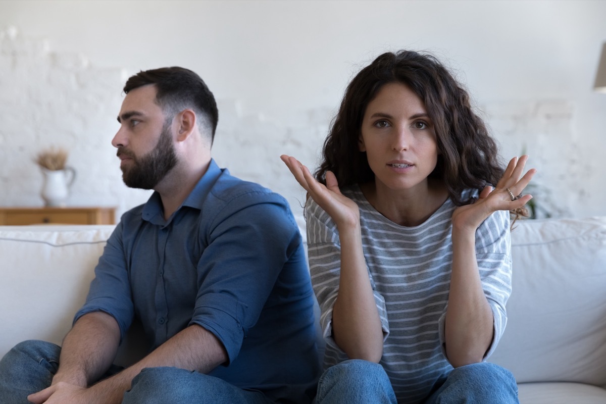 A couple on their couch; the serious wife is looking at the camera, wile a tired husband is turning away. 