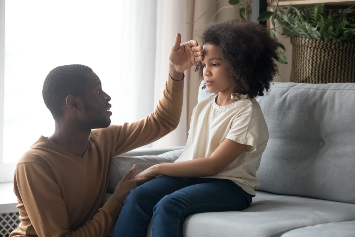 Worried dad father check temperature touch forehead of unhealthy schoolgirl daughter sitting on sofa at home