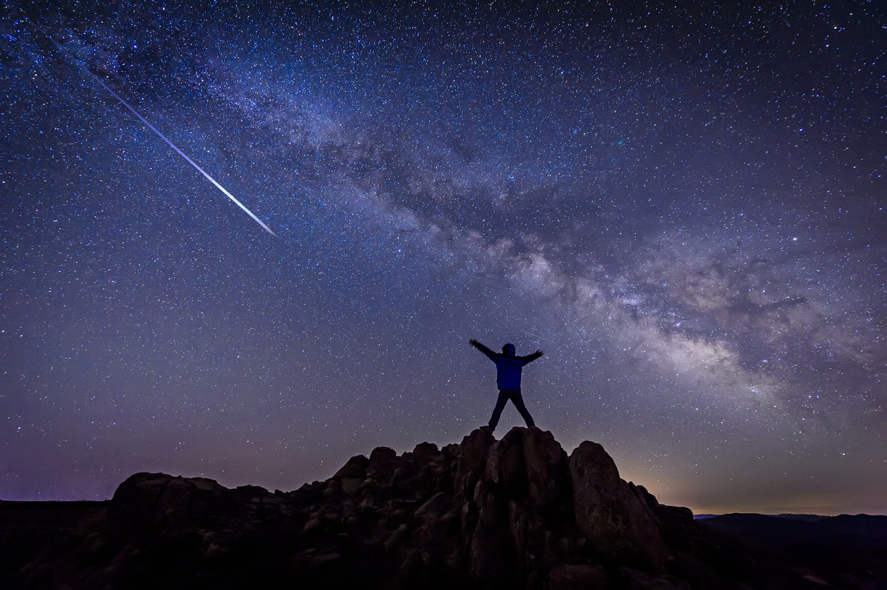 A person standing on top of a hill watching a shooting star from a meteor shower and looking at the Milky Way