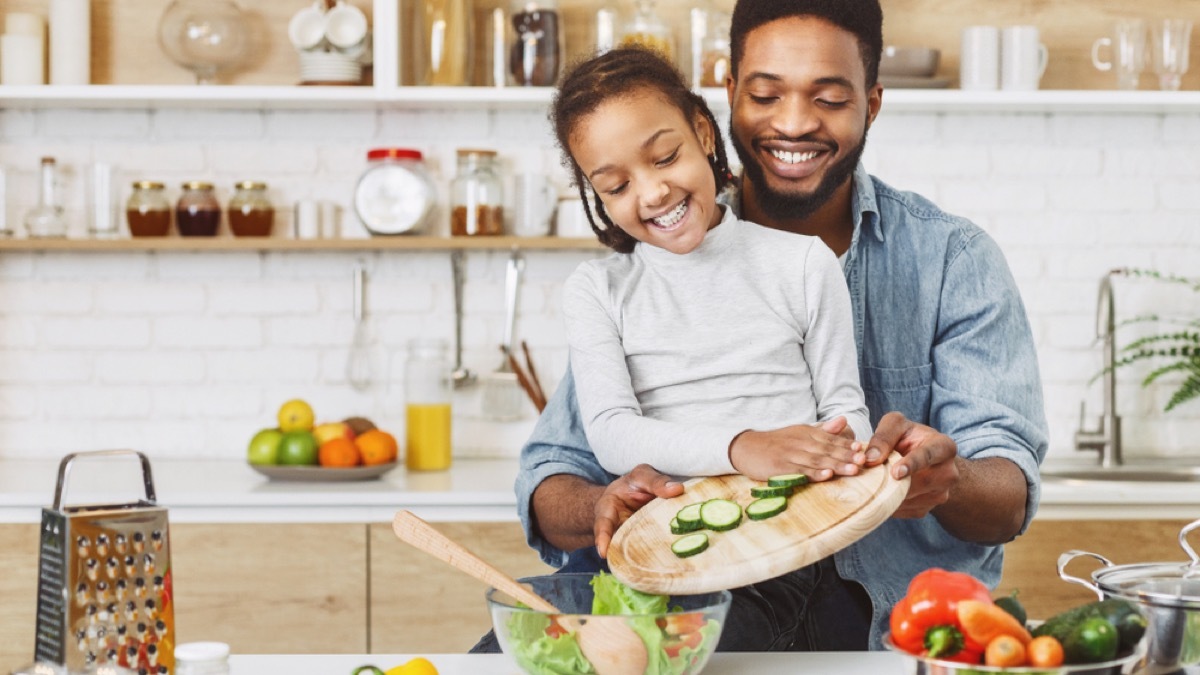 black father helping daughter push cucumbers into a salad bowl