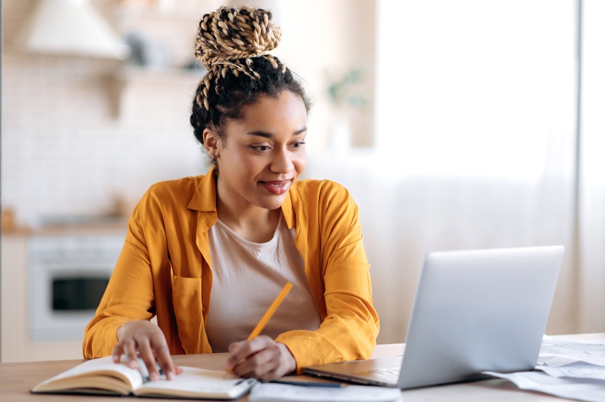 young woman wearing a yellow shirt taking notes on notepad during online lesson