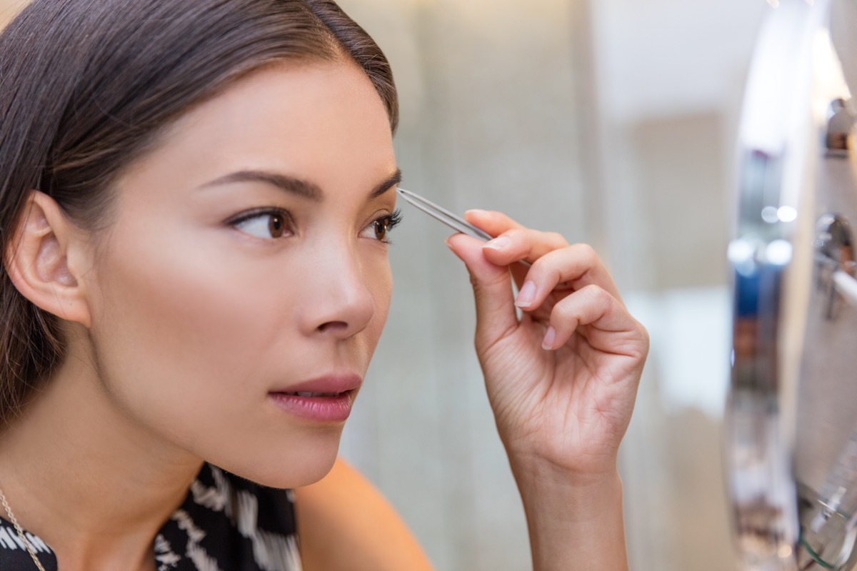 Asian woman plucking eyebrows with tweezers using eyebrow tweezer at home in bathroom makeup mirror. Closeup of a girl's face while she is removing her facial hairs. Eyebrows beauty car