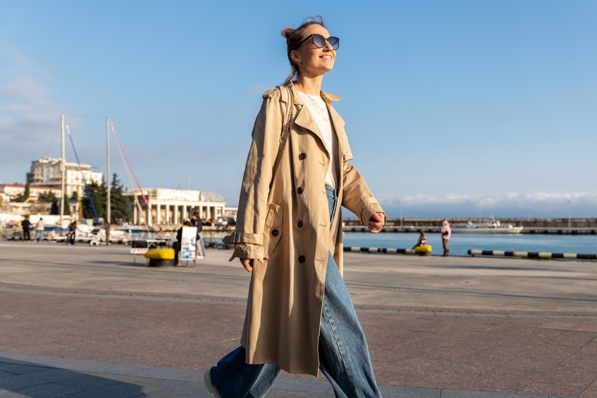 Yonug adult trendy stylish beautiful caucasian happy smiling woman enjoy walking by Yalta sea embankment on warm sunny day. Female person portrait wear jeans biege trench coat on urban city street