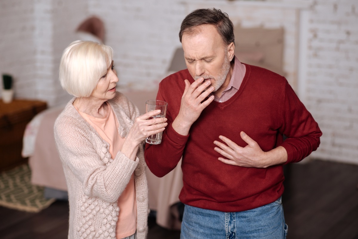 Drink some water. Elderly sick man standing and coughing strongly while his loving wife offering him water glass.