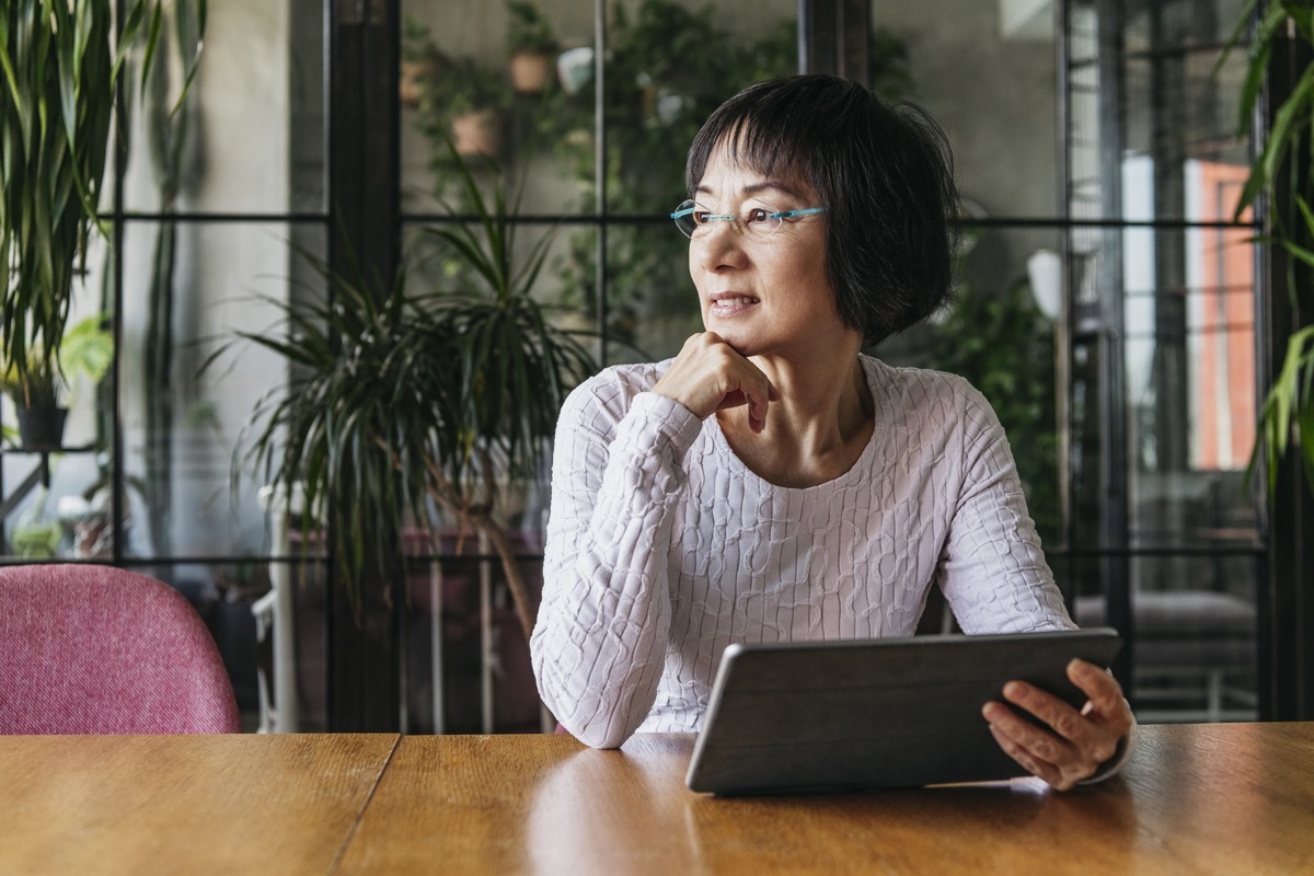 Thoughtful senior woman sitting at home with digital device, looking away and daydreaming