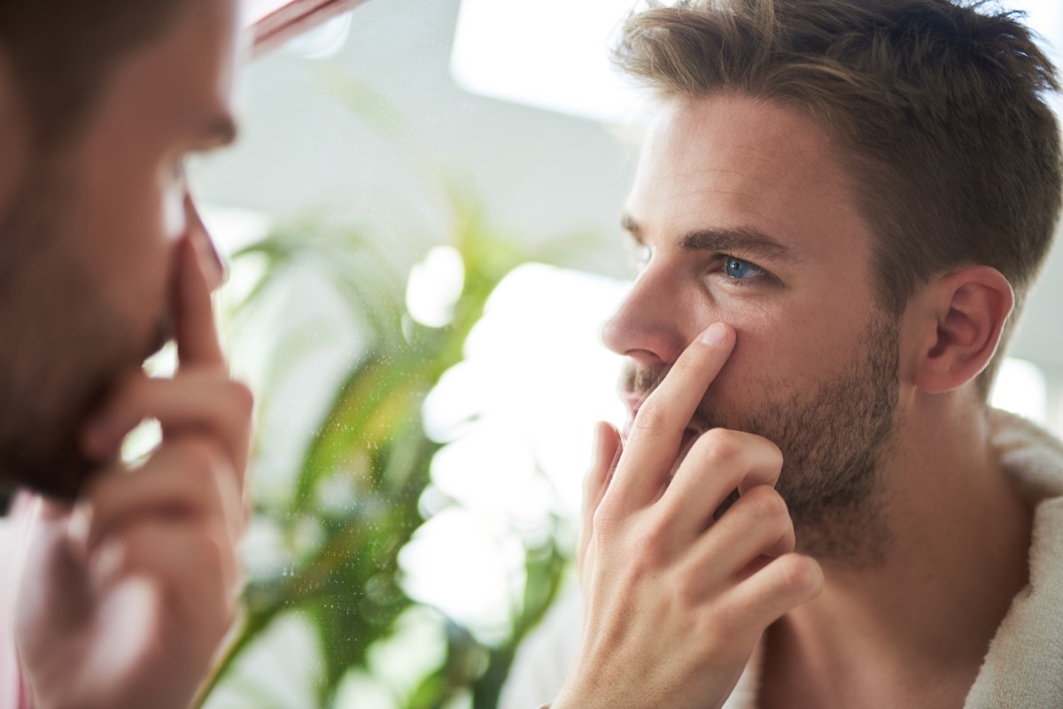 young man standing in front of mirror. He is looking precisely at face and touching skin under eyes with finger