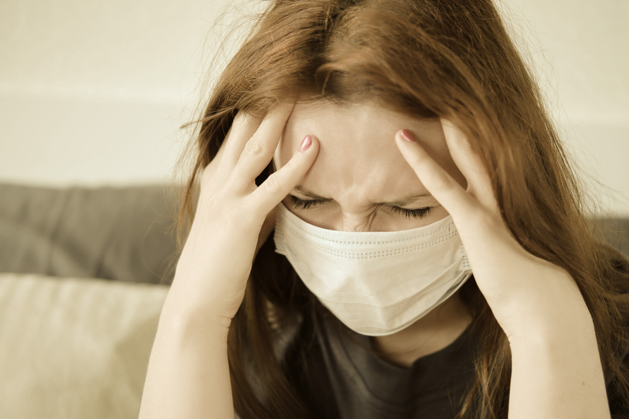 A redheaded woman in a medical mask holding her aching head