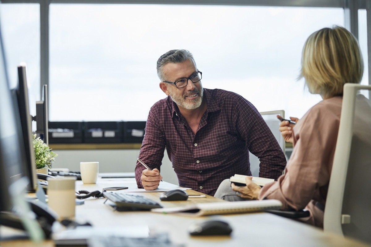 Smiling businessman communicating with colleague at desk. Male and female professionals are sitting in office. They are discussing strategies at textile industry.