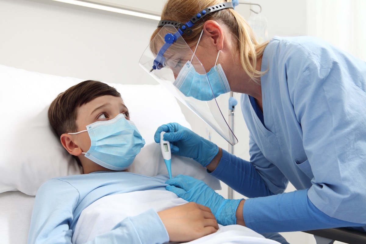 Nurse with thermometer measures fever on patient child in hospital bed, wearing protective visor and surgical mask.