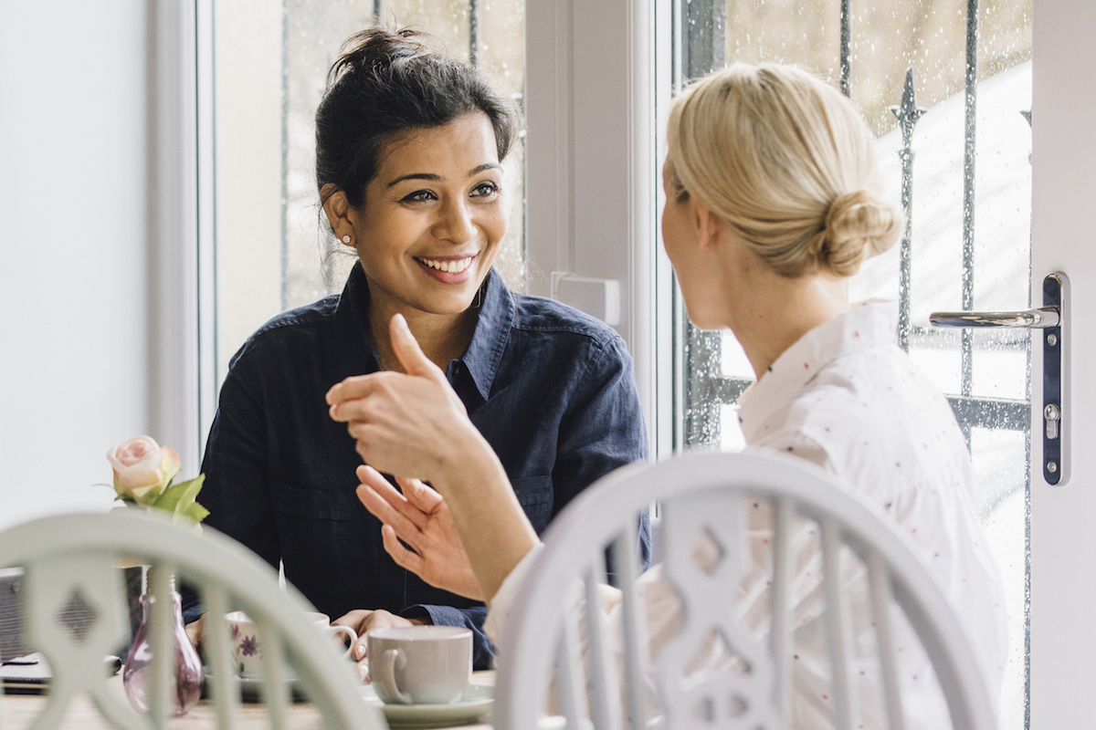 Two smiling women are sitting at a table in a cafe, socialising over tea.