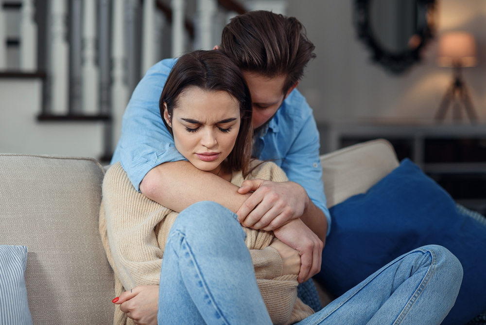 Young couple with man apologizing after fight or cheating; the woman sits on the couch while he hugs her from behind